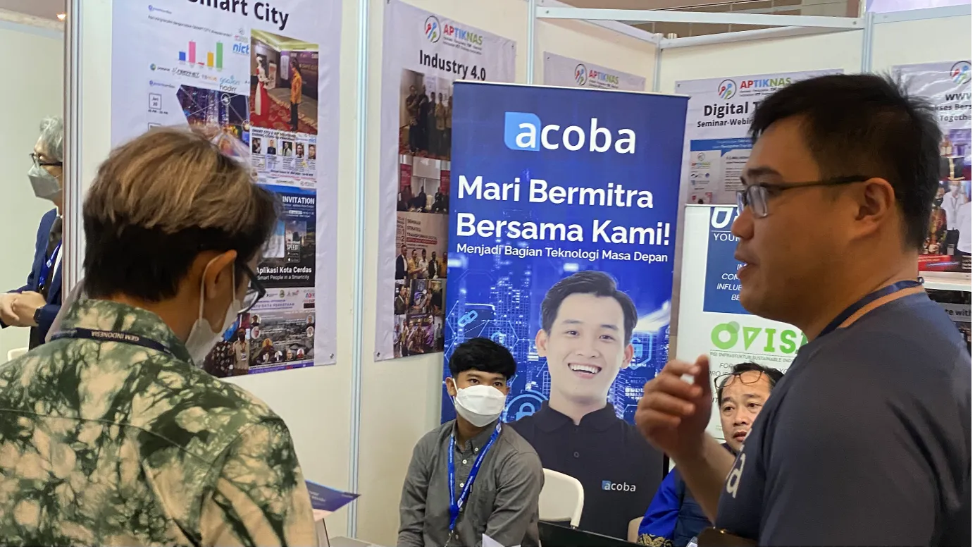 Staf Acoba menjelaskan produk kepada pengunjung di depan spanduk Mari Bermitra Bersama Kami