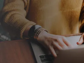 Tangan mengetik di laptop dengan buku catatan terbuka di sebelahnya