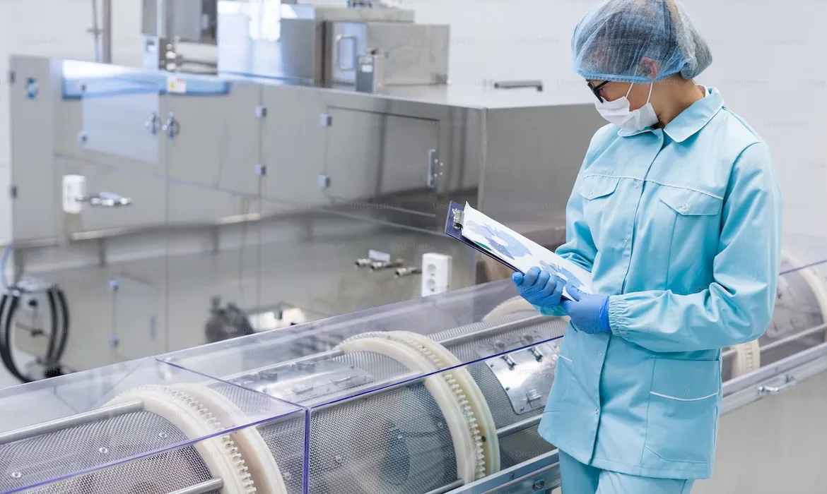 Teknisi berpakaian steril lengkap memeriksa catatan di samping peralatan produksi laboratorium